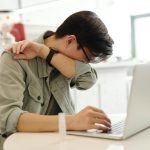 A man sneezes into his elbow while using a laptop, depicting common cold symptoms in a modern workspace.